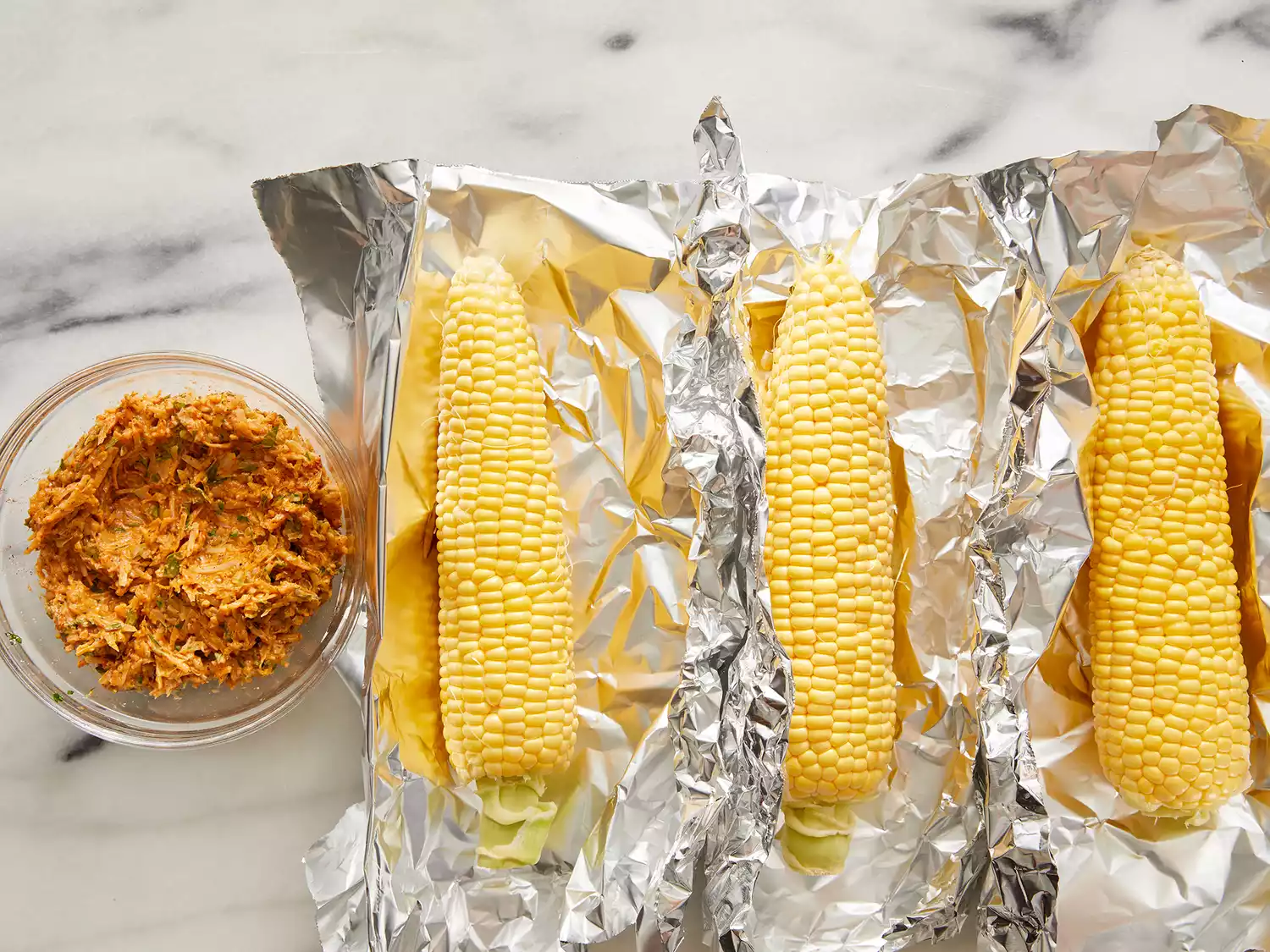 Ears of corn placed in aluminum foil and mayonnaise, parmesan cheese, chili powder, black pepper and parsley mixed together in a bowl.