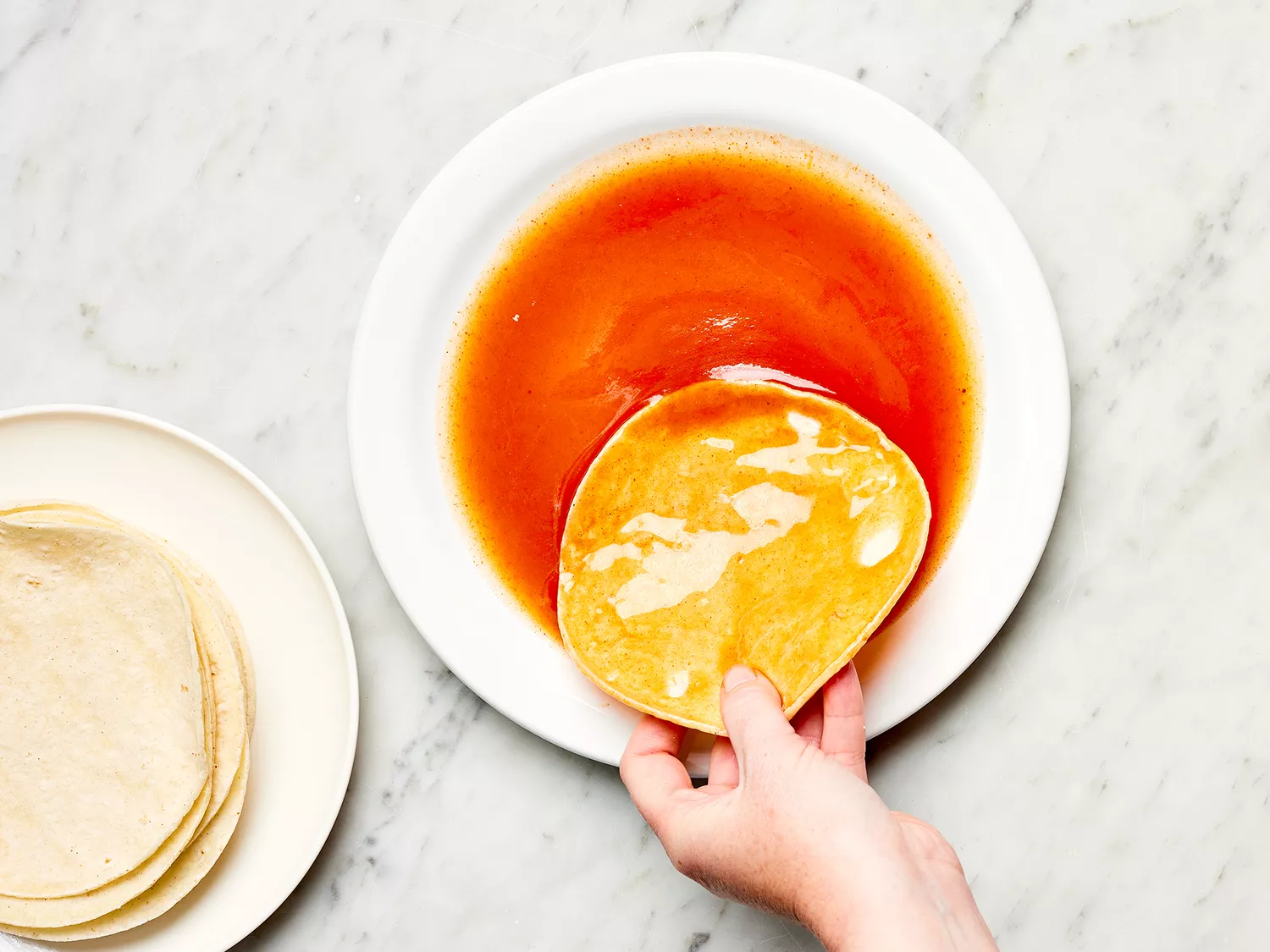 Hand dipping a corn tortilla into red enchilada sauce on a white plate 