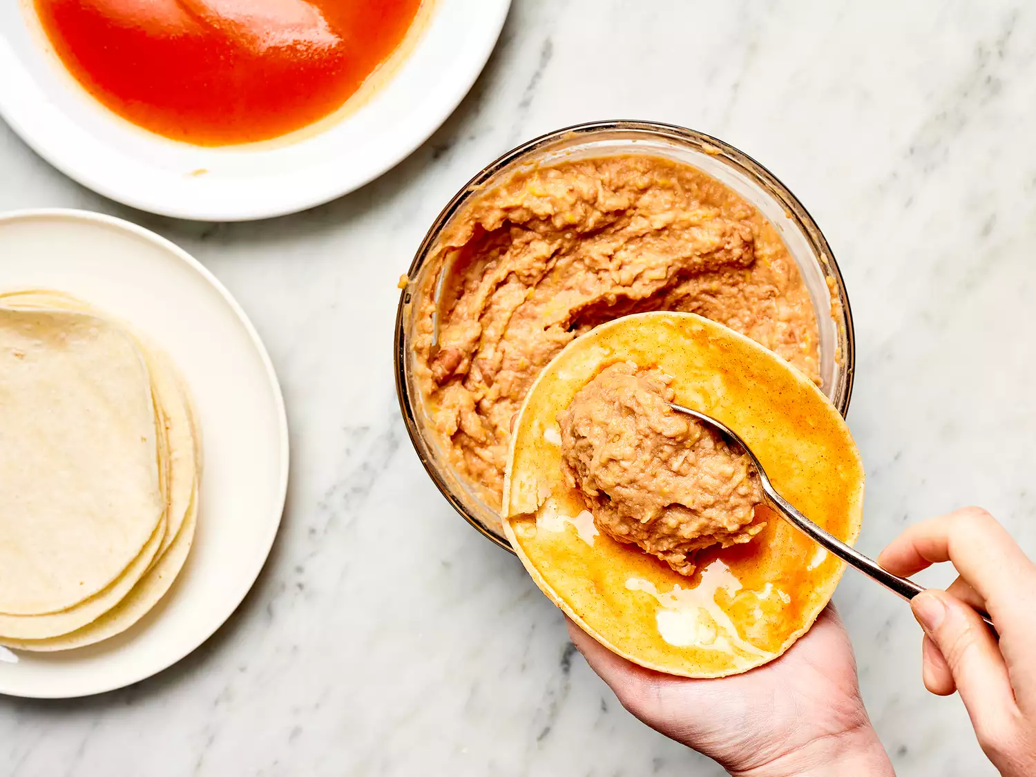 Hand spooning refried beans onto a corn tortilla covered with red enchilada sauce