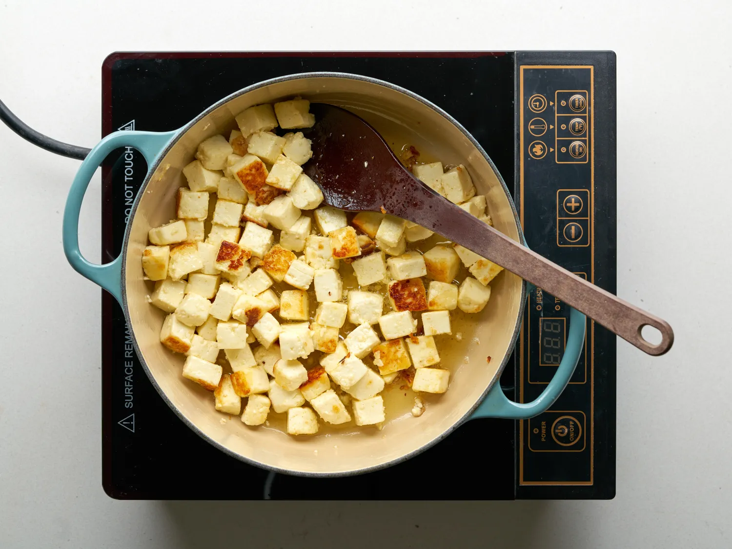 Paneer cubes cooking in a pan on an induction stove with a wooden spoon