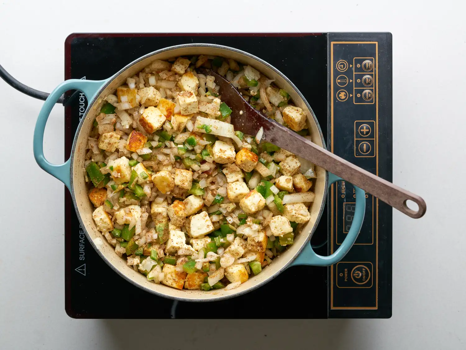 Cooking paneer tikka masala in a pot on an electric stove, paneer cubes and vegetables being stirred with a wooden spatula