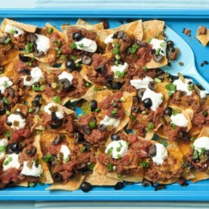 Overhead view of a tray of super nacos recipe with tortilla chips, ground beef, black olives, salsa, green onions, cheese, and sour cream toppings