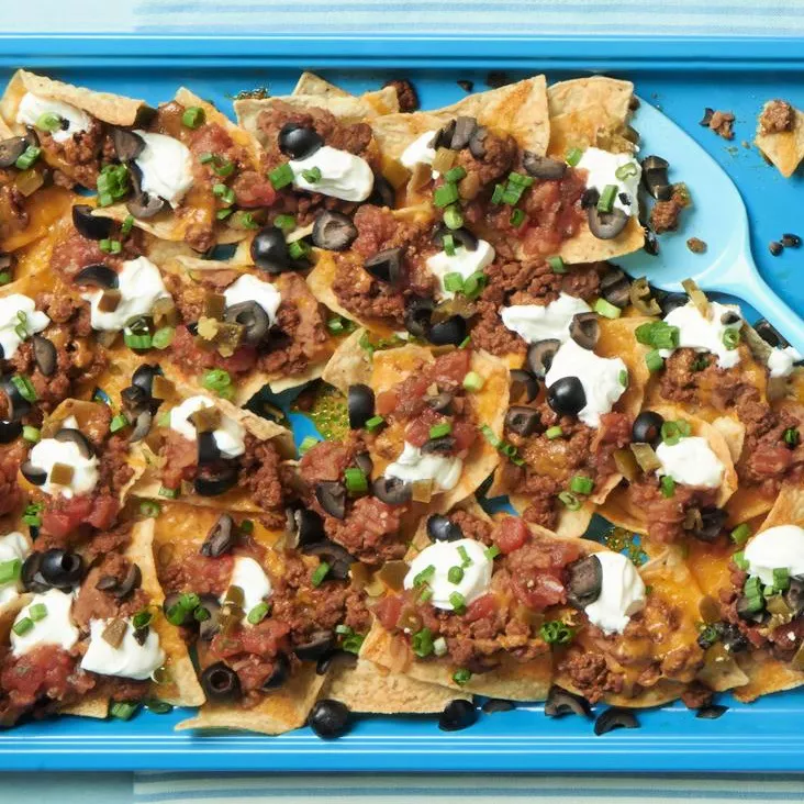 Overhead view of a tray of super nacos recipe with tortilla chips, ground beef, black olives, salsa, green onions, cheese, and sour cream toppings