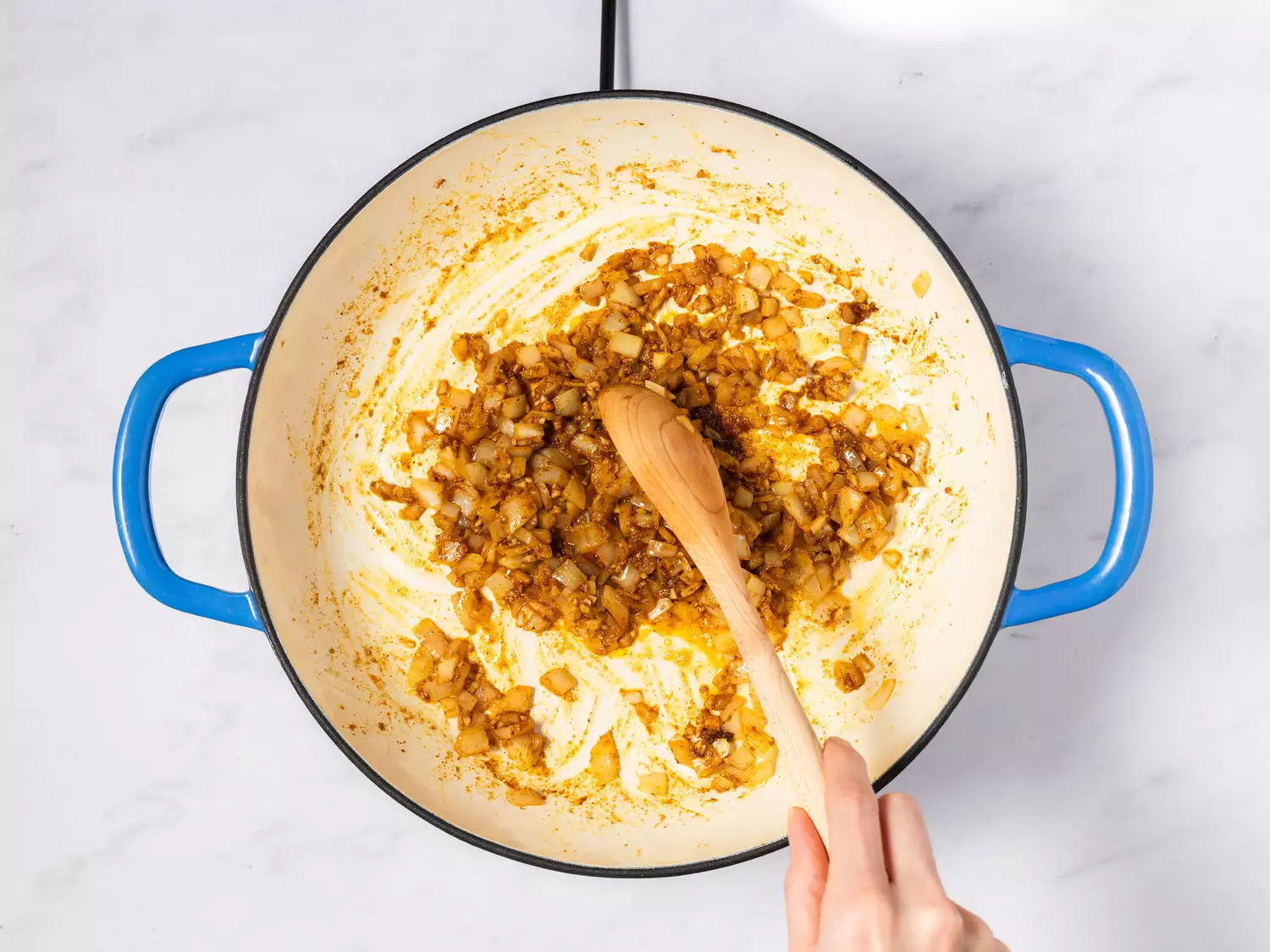 A hand stirring sautéed onions and spices in a pot with a wooden spoon