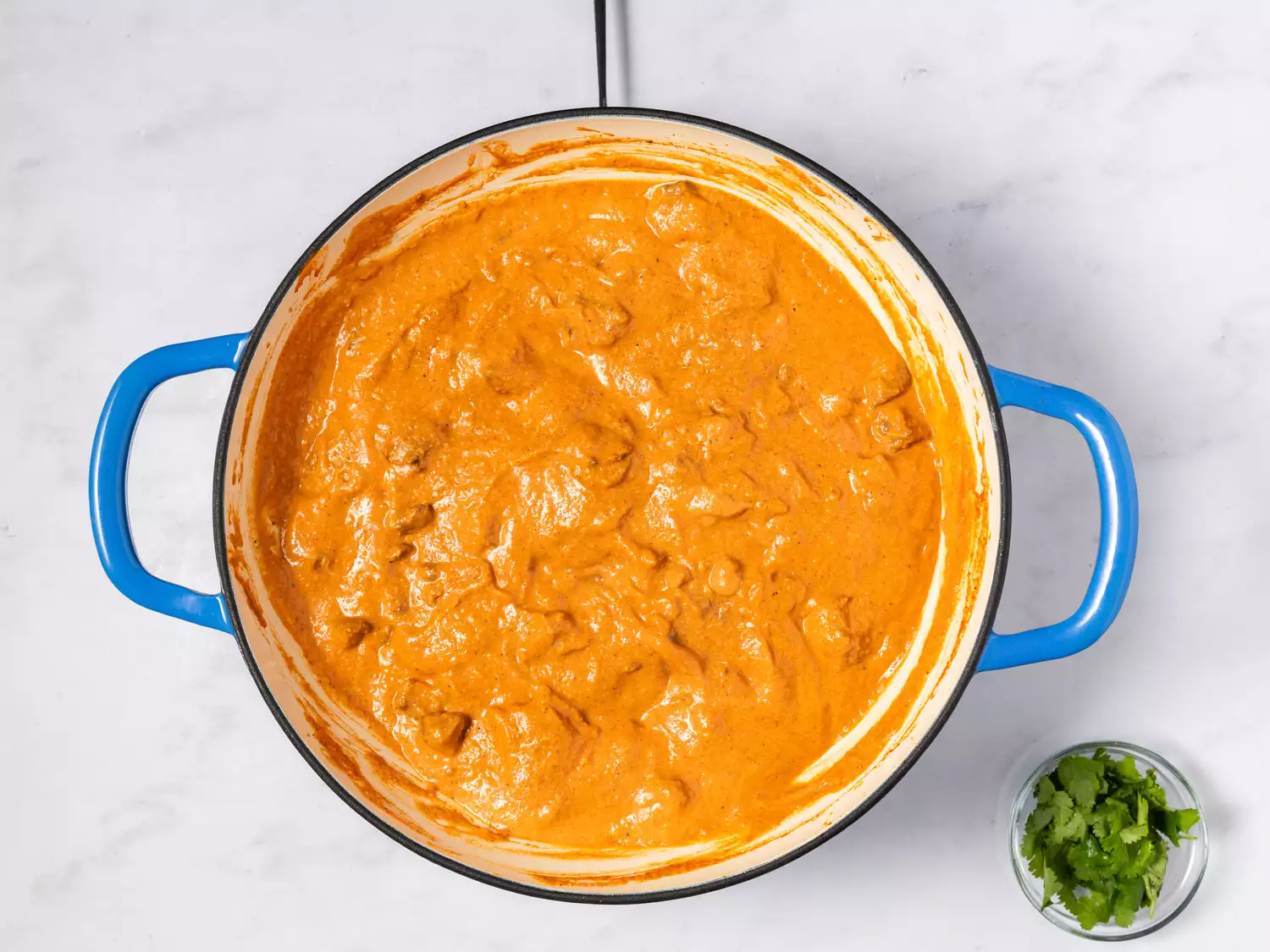 A pot of butter chicken simmering, garnished with a side of chopped herbs