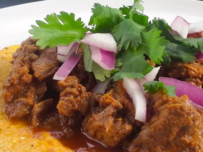 Close up view slow cooker beef barbacoa over grits, garnished red onions and cilantro, on a white plate