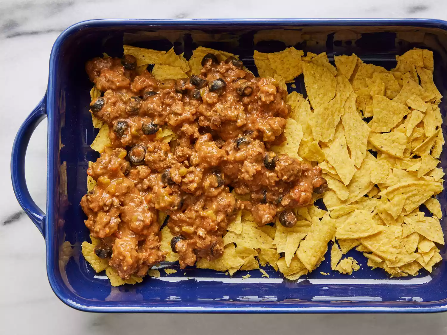 A blue casserole dish with broken taco shells on the bottom, halfway covered with cooked taco meat. 