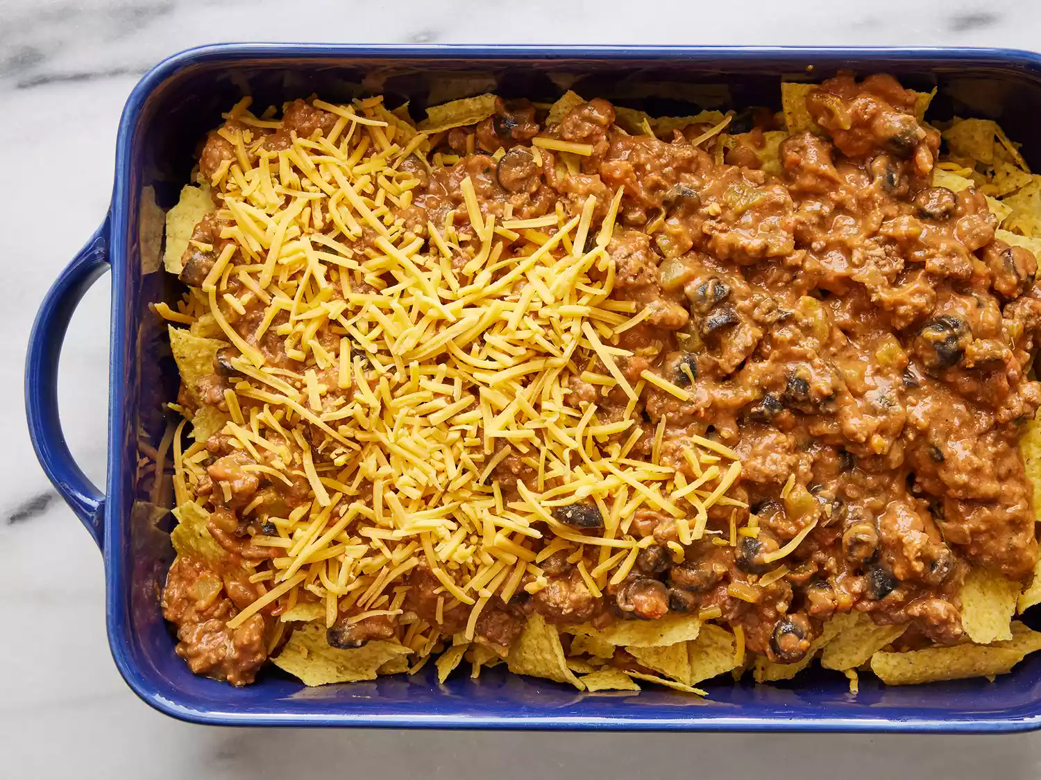 A blue casserole dish with cheese sprinkled over taco meat and broken taco shells. 