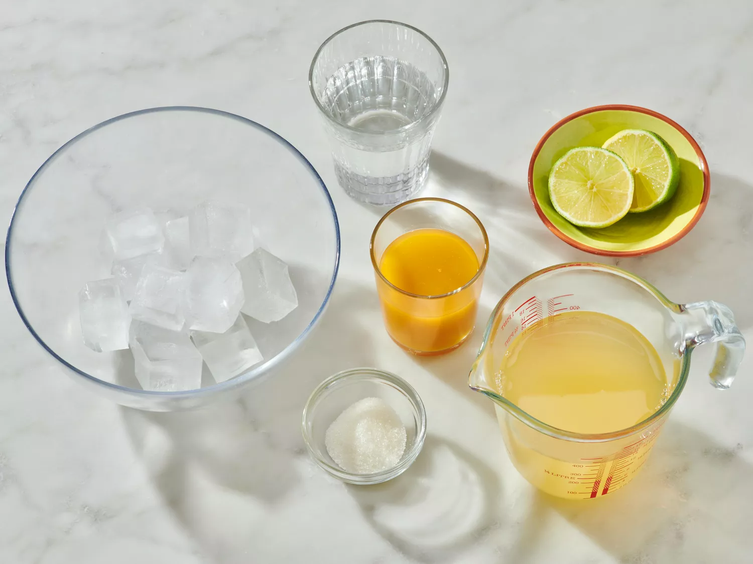 Ingredients for mango margaritas including a bowl of ice, lime slices, juice, and other components arranged on a light surface
