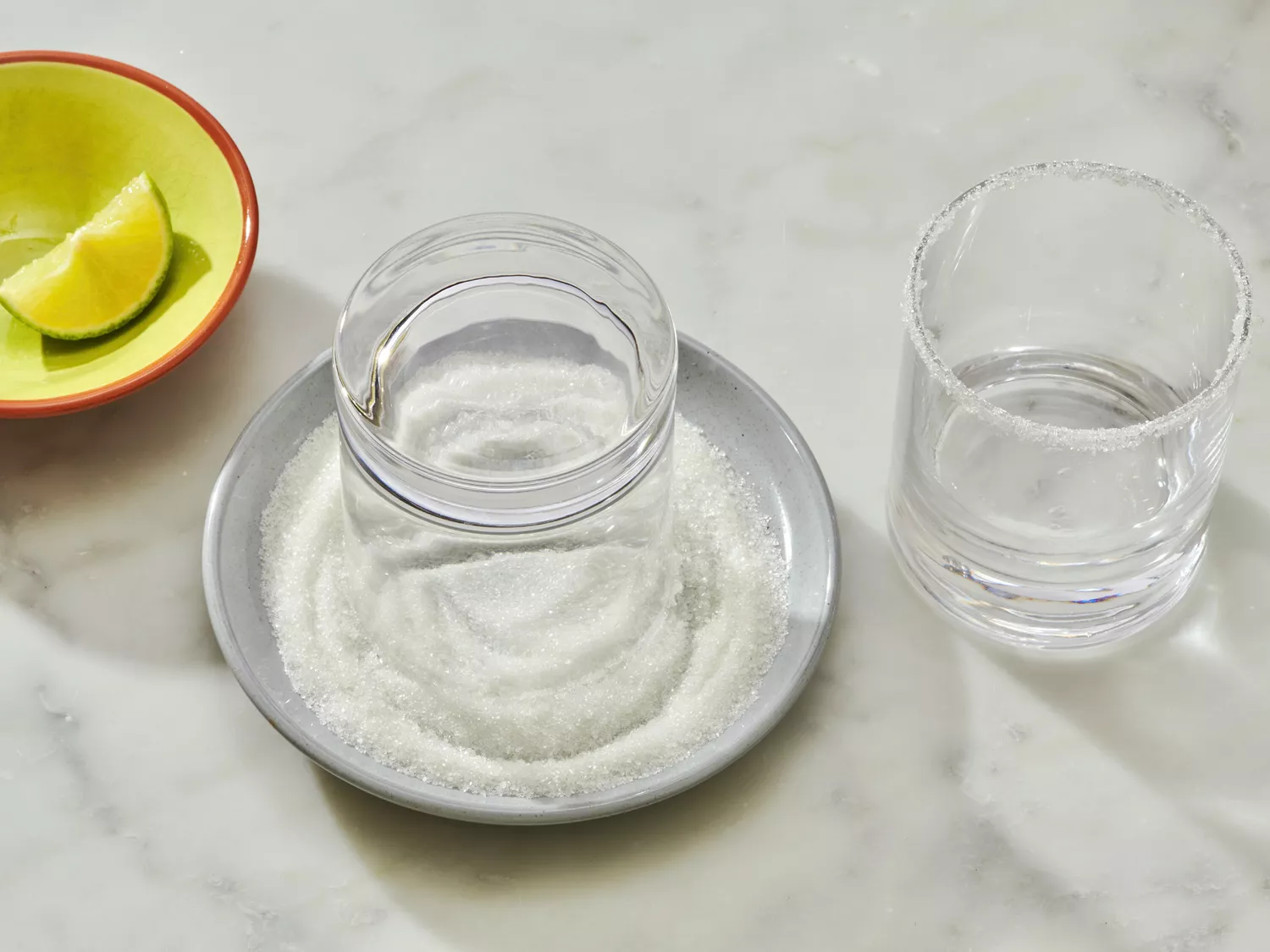 Glass rim being coated with salt on a plate next to a salted glass and a lime wedge in a dish