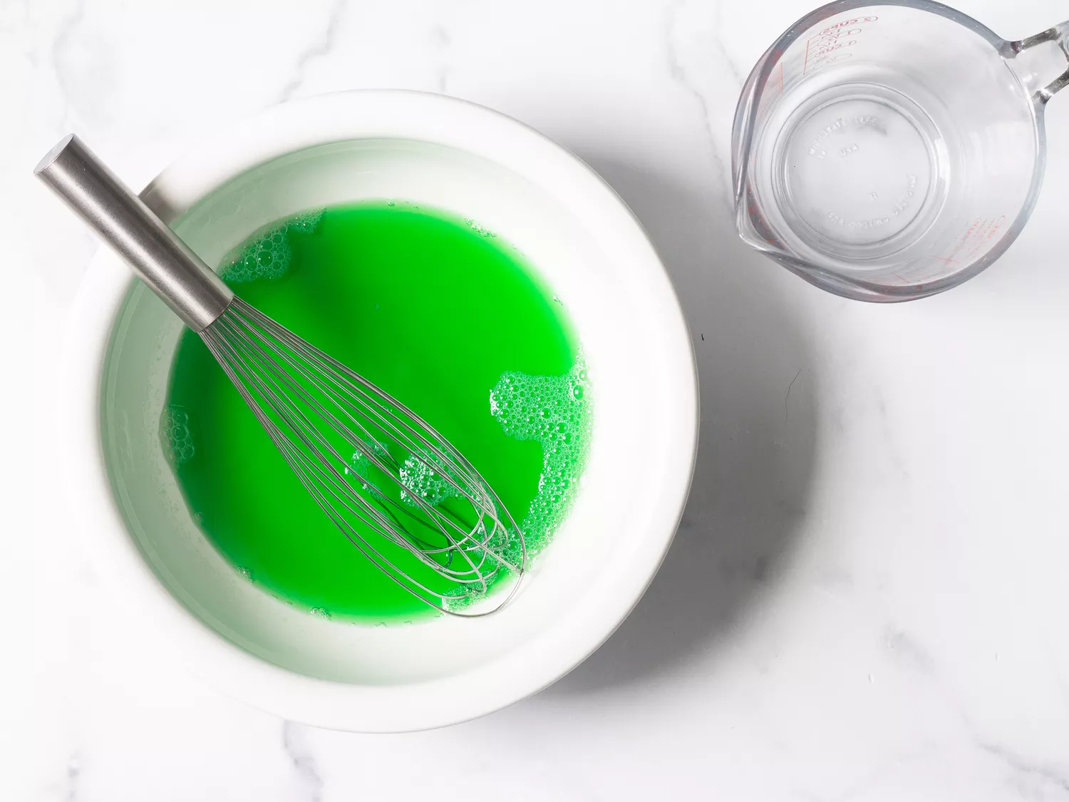 Whisk in mixture of green liquid in a white bowl, with a measuring cup nearby