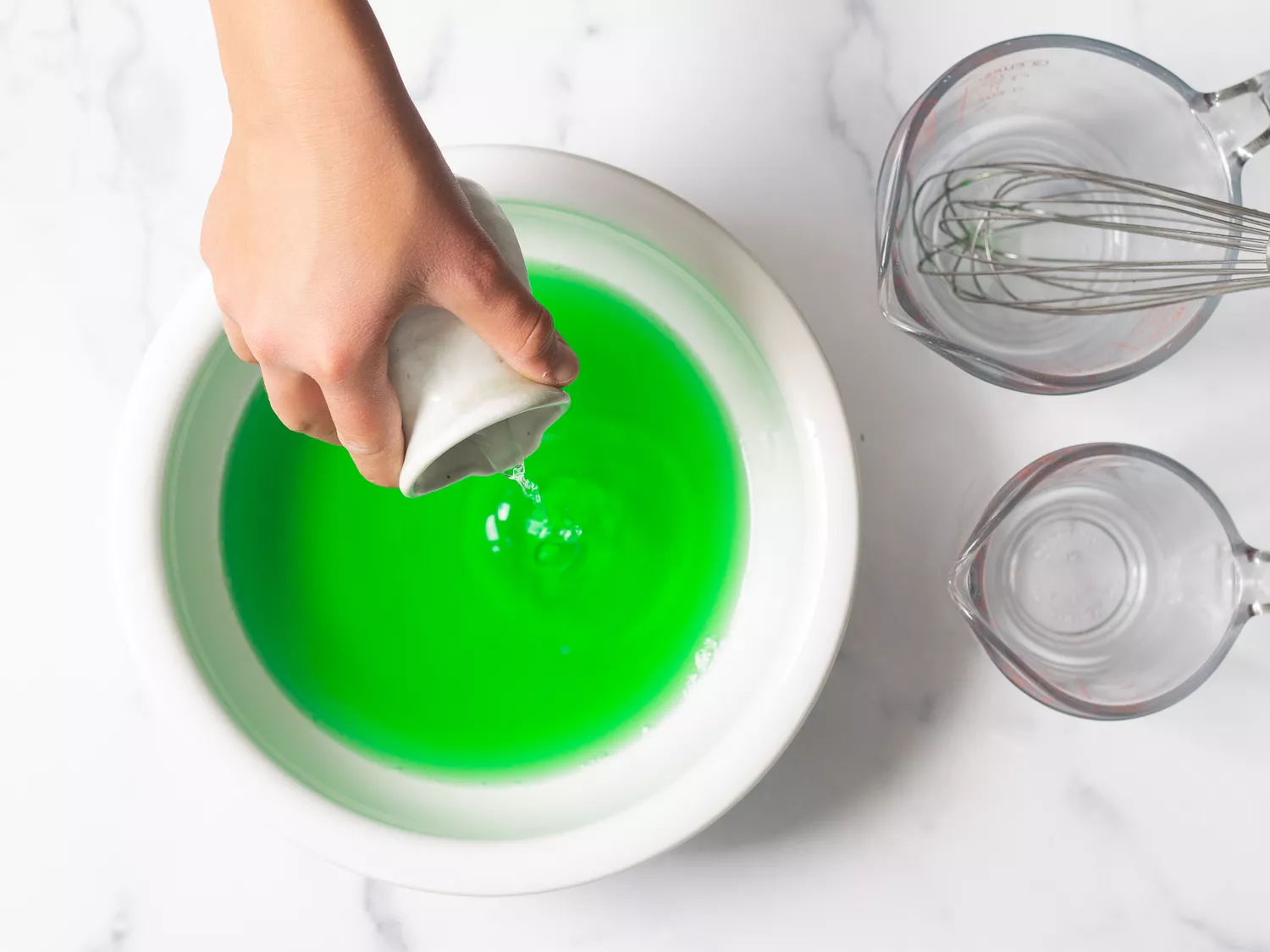 A hand pouring liquid into a bowl containing green gelatin mixture, with measuring cups and a whisk nearby