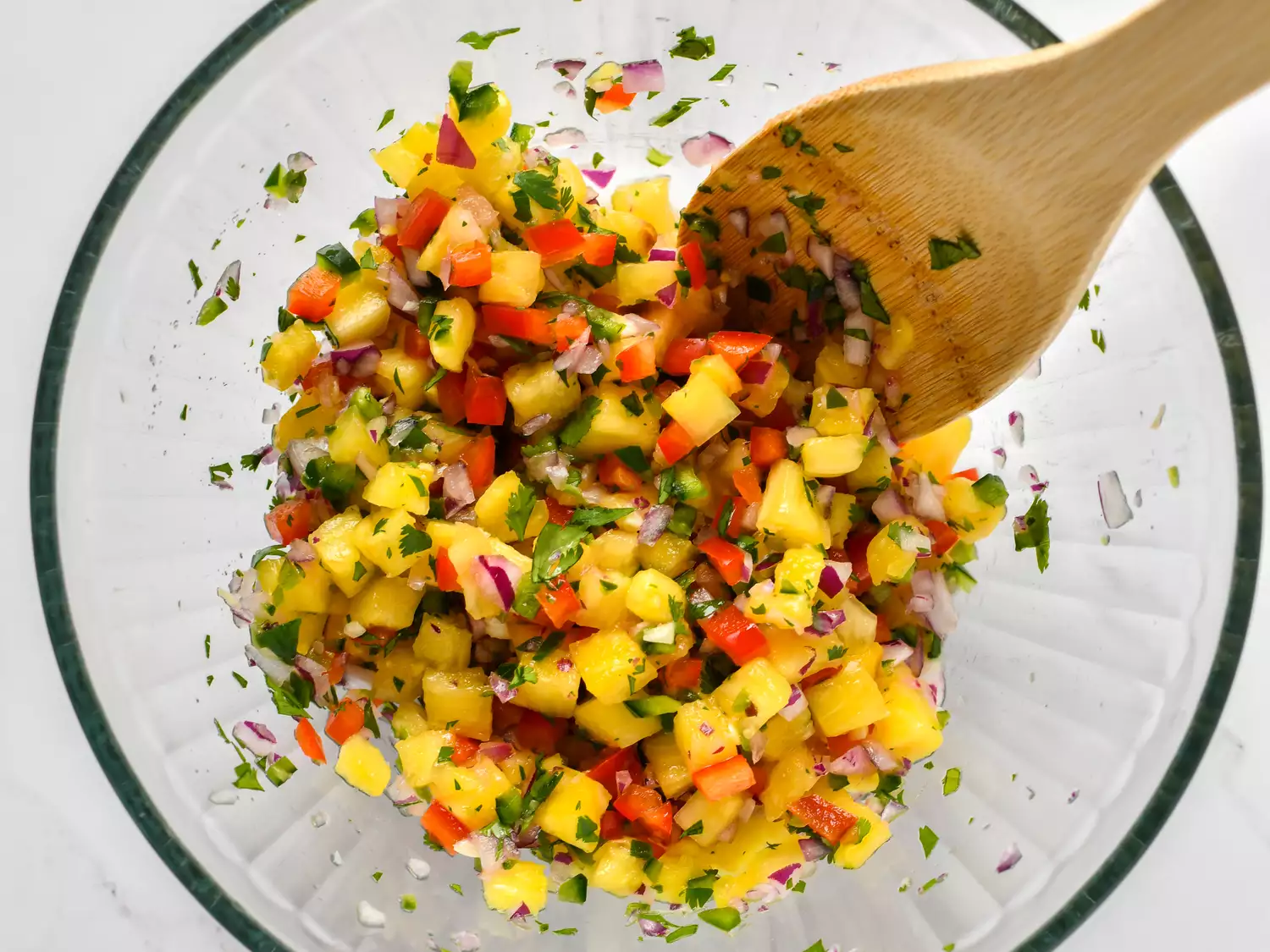 A glass bowl filled with diced pineapple and other ingredients for salsa, stirred with a wooden spoon