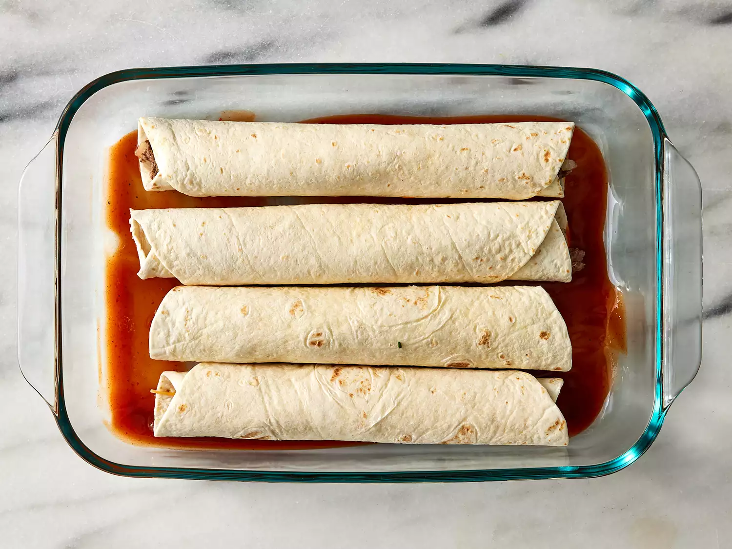 beef enchiladas placed seam side down in a baking dish with enchilada sauce in the bottom.