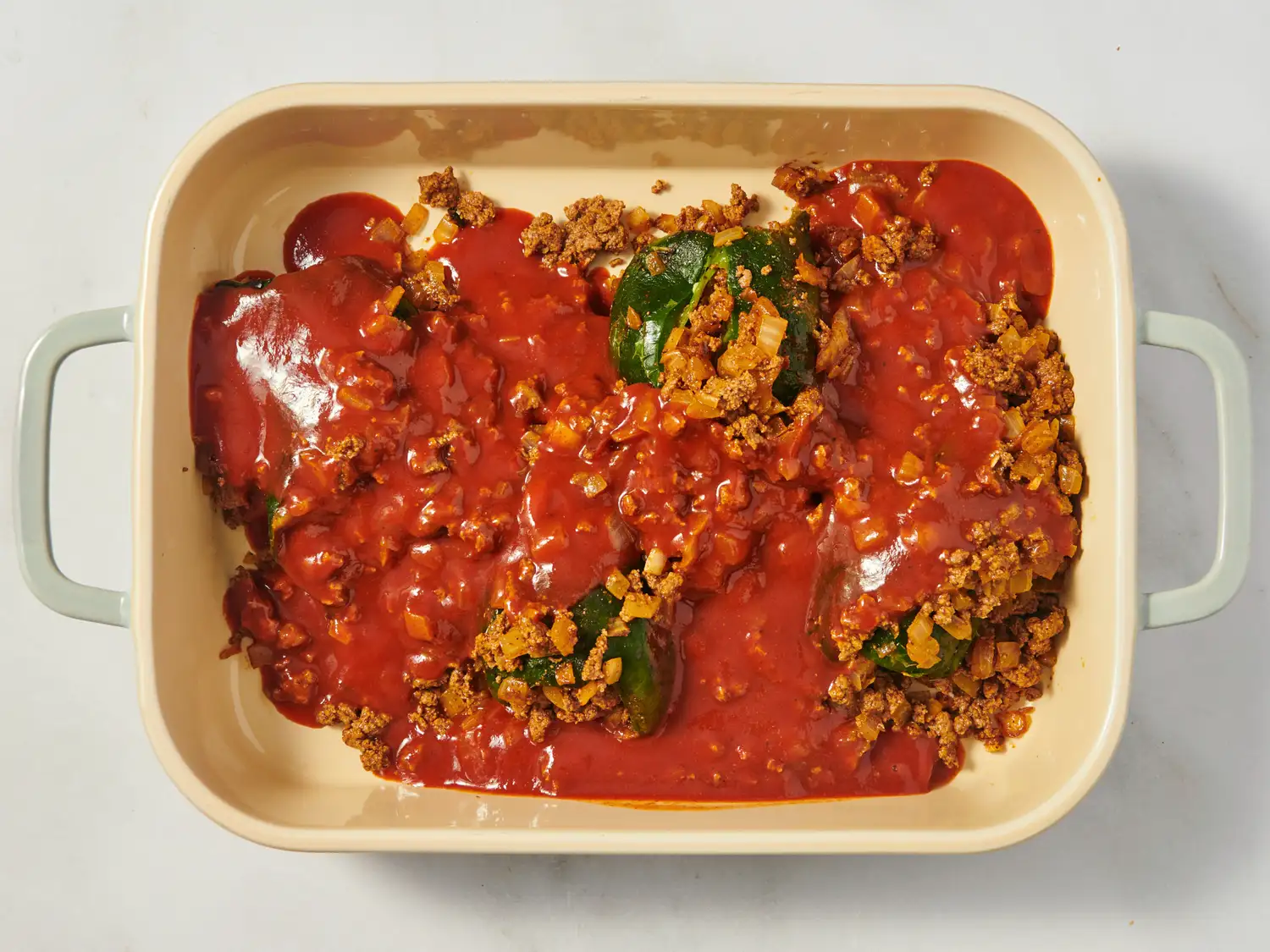 Baking dish filled with stuffed poblano peppers topped with a tomatobased sauce and ground meat