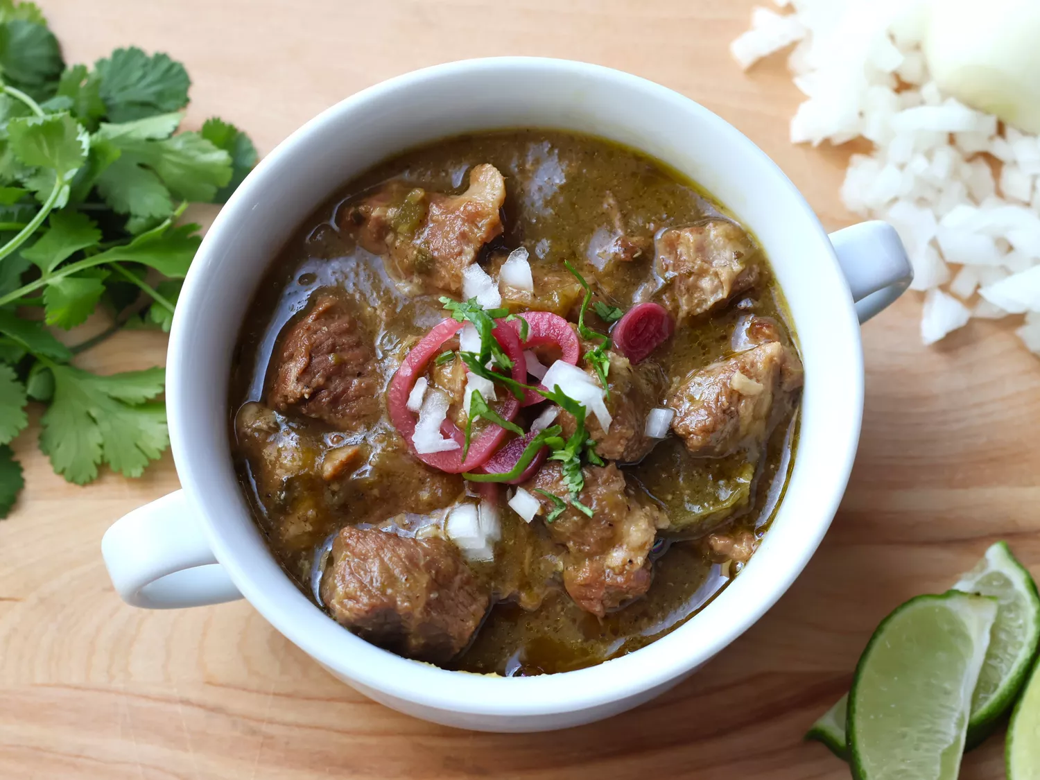 Pork chili in white bowl garnished with onions and cilantro and lime slices on the side