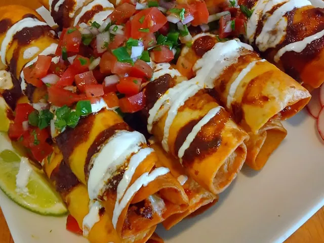 A close up look at a pile of golden spicy chicken taquitos topped with sour cream and fresh pico de gallo.