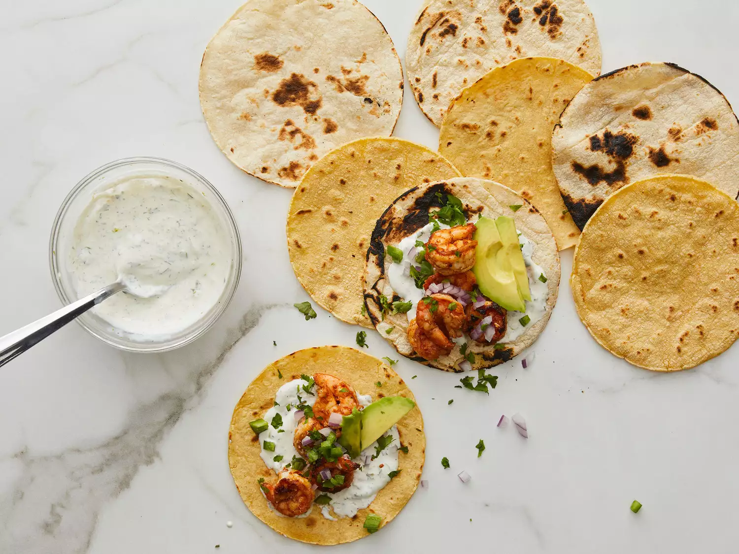 Overhead of tortillas being filled with shrimp, crema, and cilantro with a bowl of crema off to the side. 