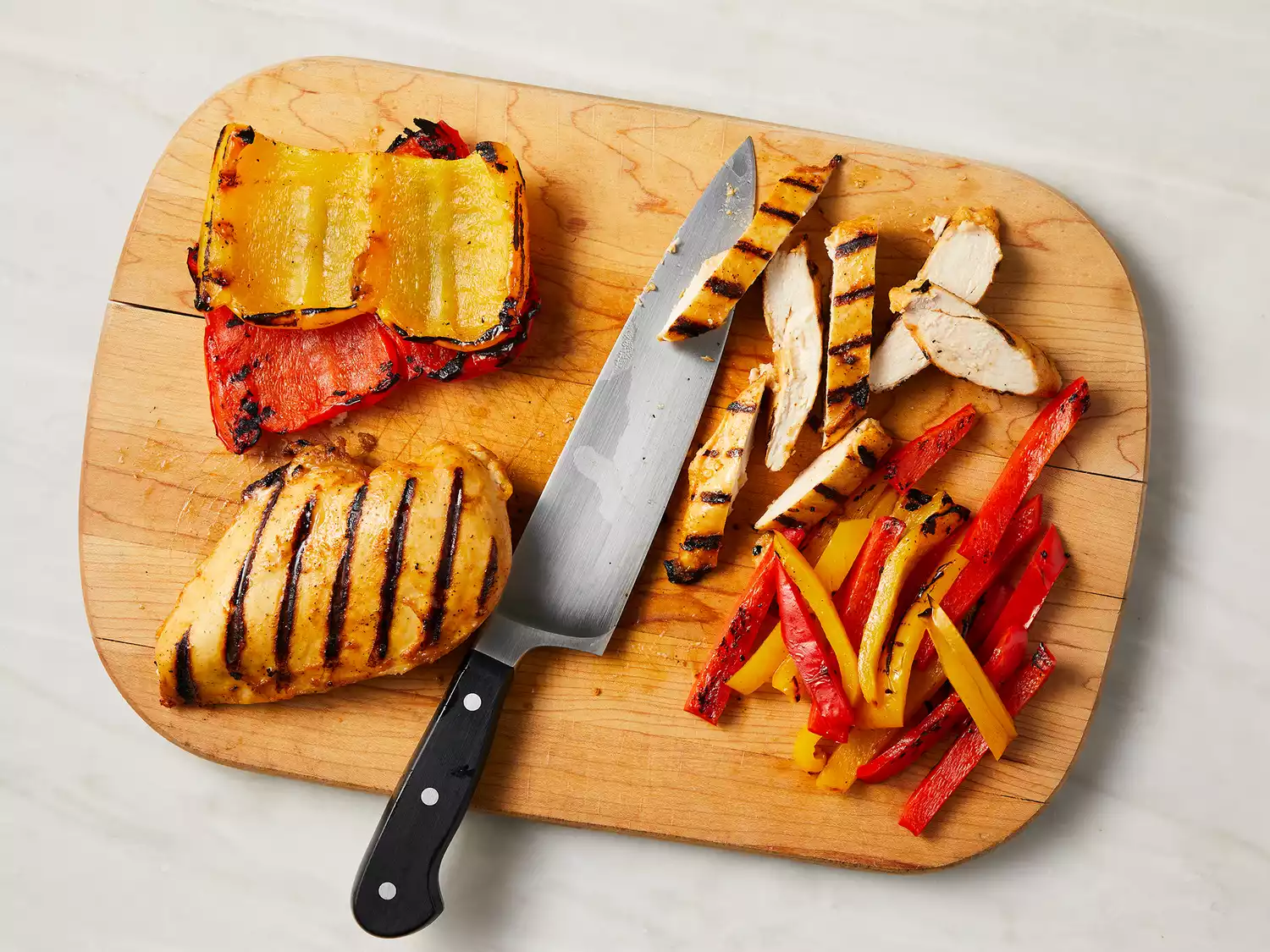 Overhead of chicken and bell peppers being sliced into strips on a cutting board. 