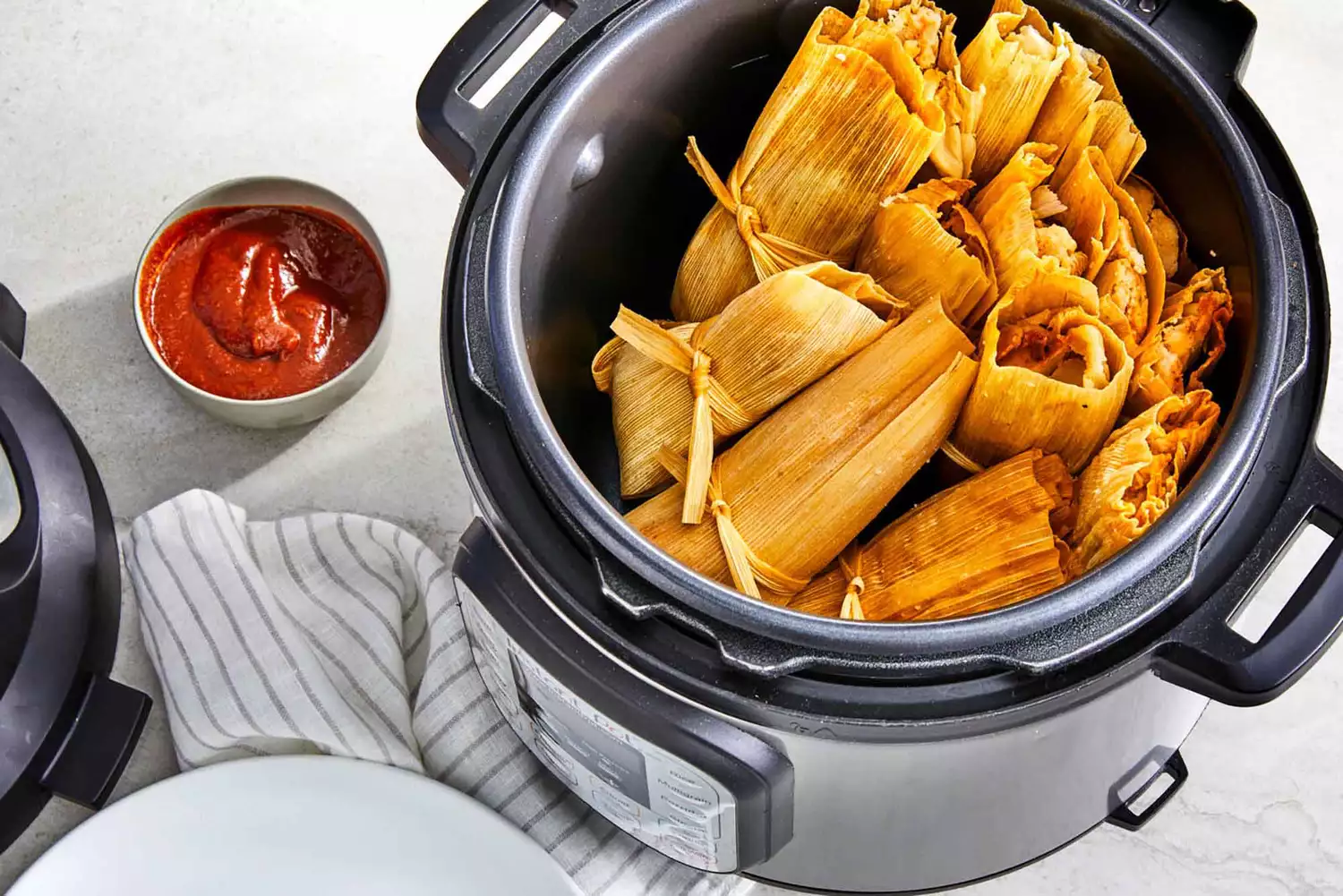 A high angle view of instant pot tamales in an instant pot with a red pepper sauce in a small bowl to the side.