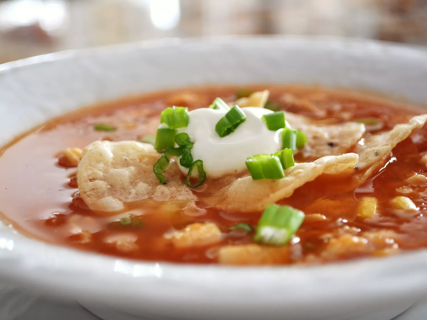 Closeup side view of a bowl of chicken tortilla soup garnished with tortilla chips, a dollop of sour cream, and chopped green onions
