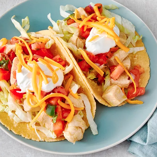Two chicken tacos on corn tortillas, topped with onions, tomatoes, sour cream and shredded cheddar