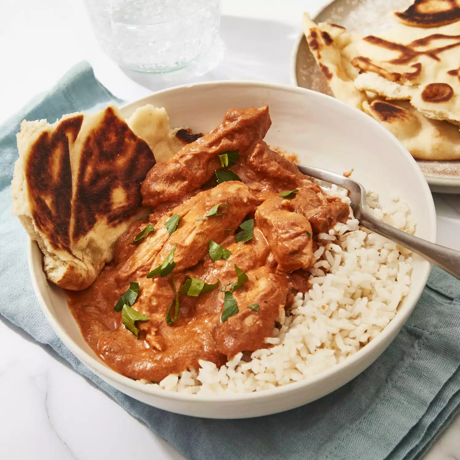 A white bowl sits on a teal linen with chicken korma. served over rice and with naan bread
