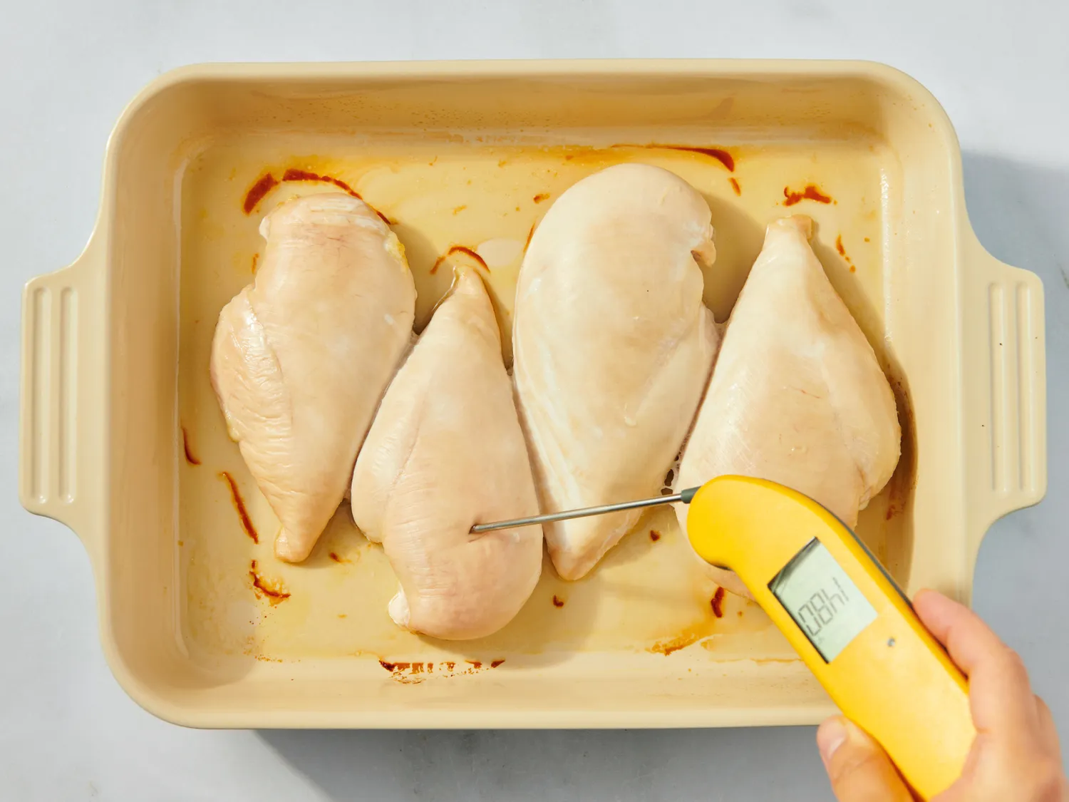 Four cooked chicken breasts on a baking dish a hand holding a thermometer inserted into one of them