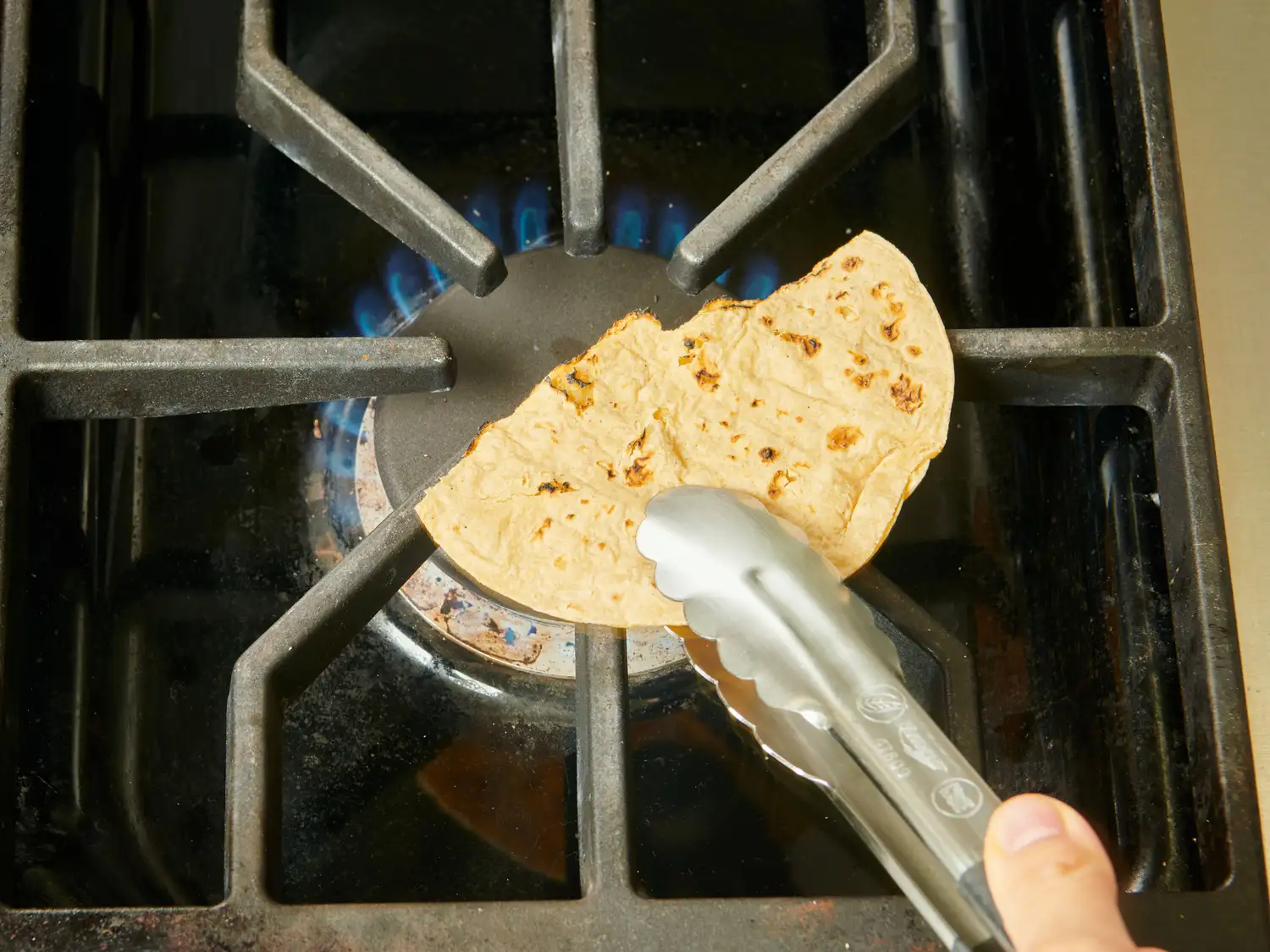 A tortilla being heated on a stovetop burner using tongs