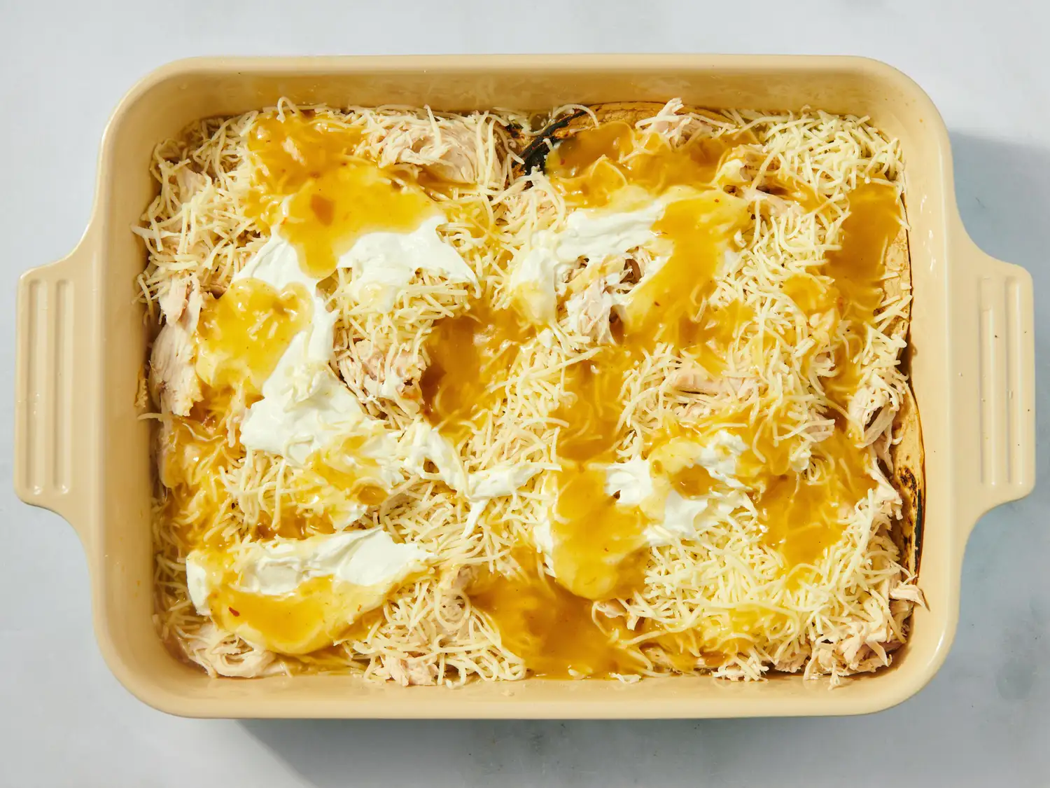 A casserole dish with shredded cheese chicken green chili sauce and sour cream layered as part of enchilada casserole preparation