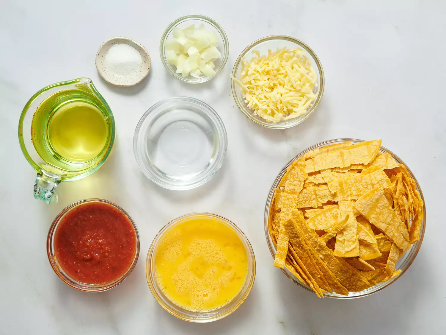 Ingredients in bowls and plates, including tortilla chips, cheese, salsa, eggs, and chopped onions, arranged on a white surface