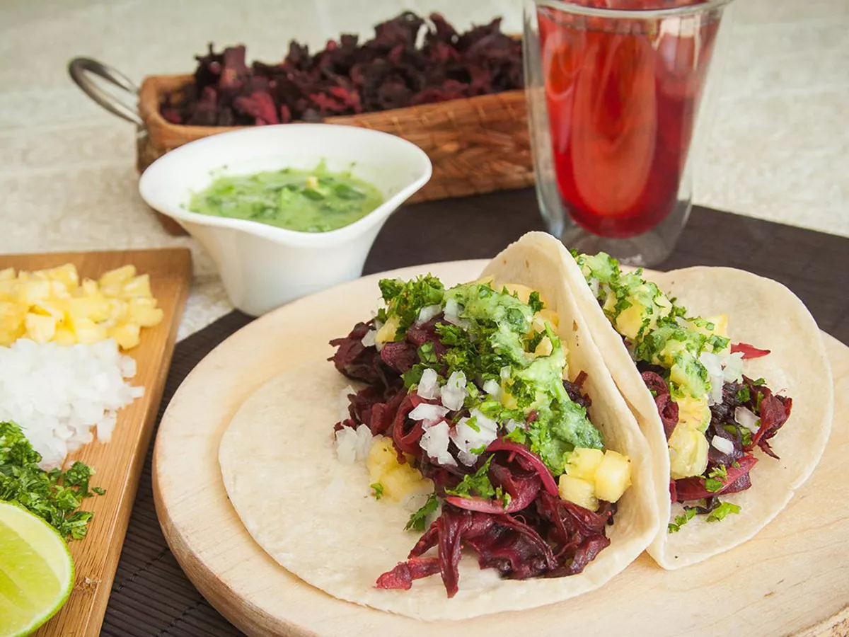 Close up view of tacos de jamaica (vegan hibiscus tacos) serve with green sauce in a sauce cup and diced herbs