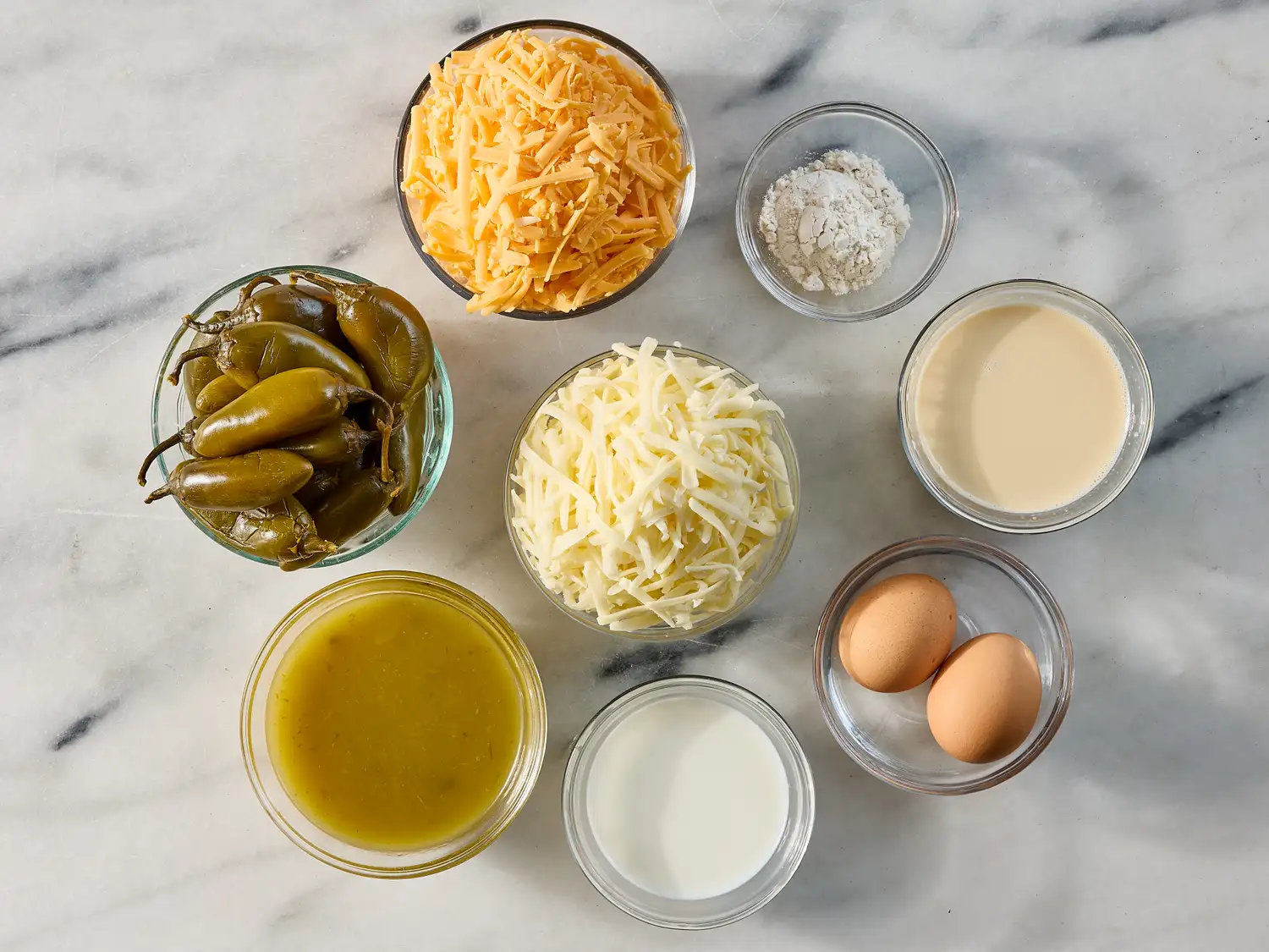 Ingredients for a chili relleno casserole including shredded cheeses canned peppers eggs milk and seasonings on a marble surface