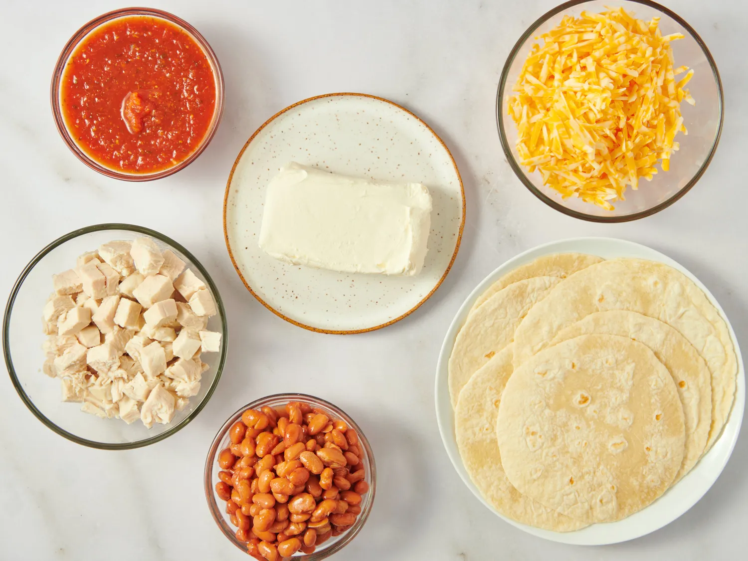 Ingredients for chicken enchiladas including tortillas cheese beans sauce chicken and a white block of cream cheese