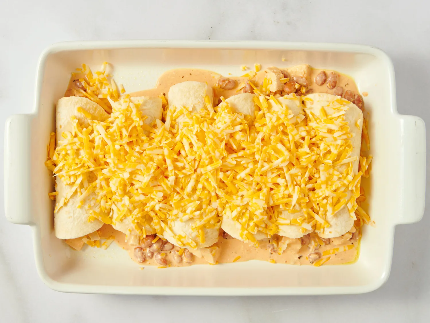 Baking dish with filled tortillas topped with shredded cheese prepared for enchiladas