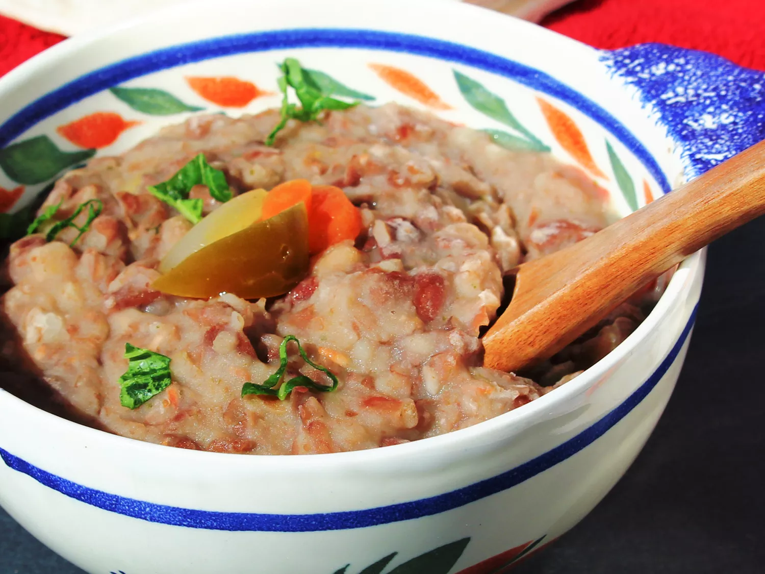 Close up view of vegetarian refried beans garnished with peppers, carrots and fresh herbs in a bowl with a wooden spoon