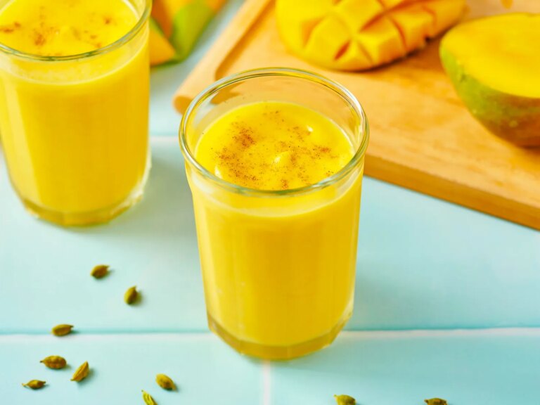 Two glasses of restaurant style mango lassi on a table with cut mangoes and cardamom pods