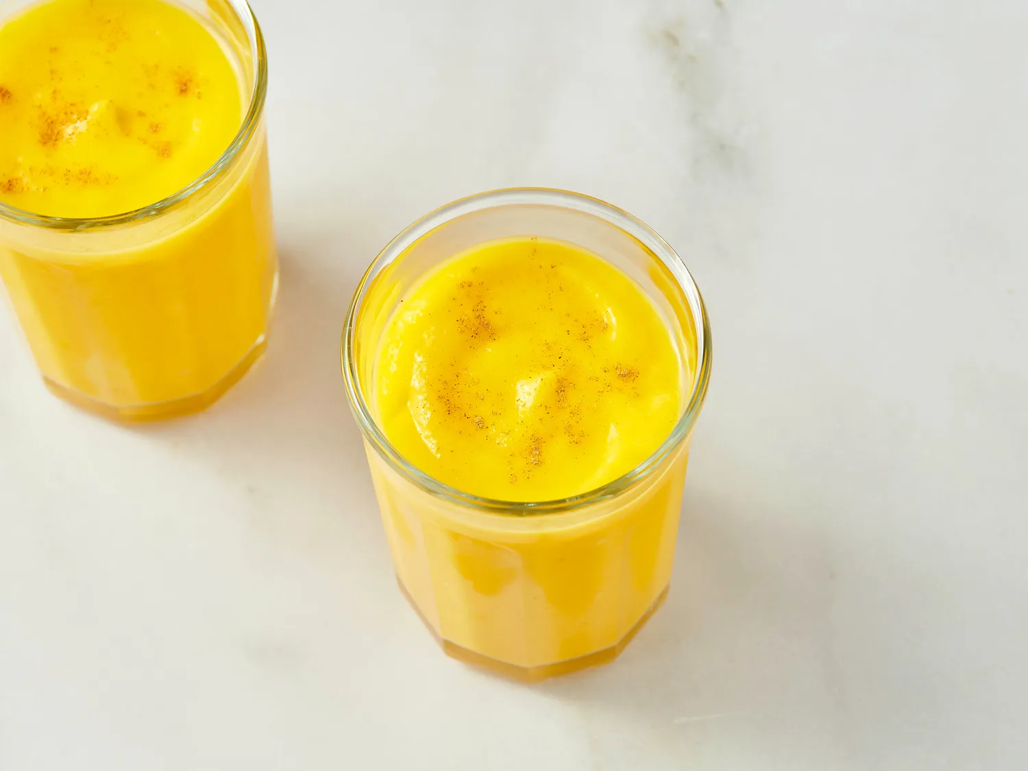 Two glasses of mango lassi on a marble surface, garnished with a sprinkle of spice