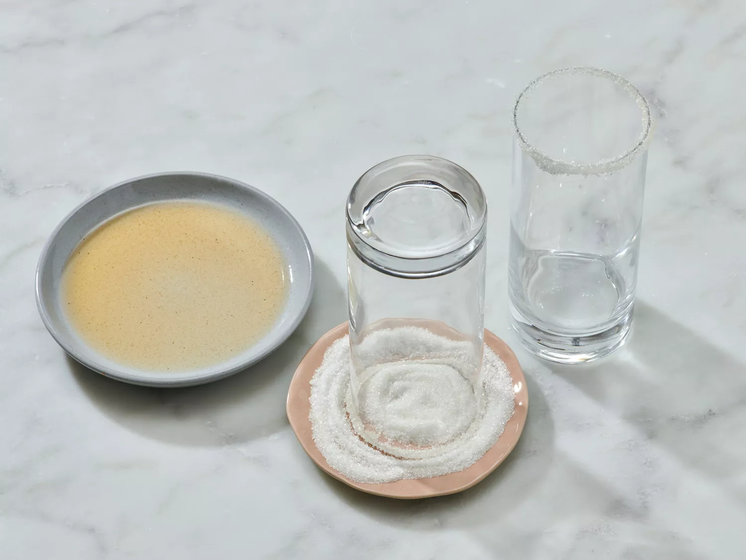 A glass being prepared by dipping its rim into salt on a plate, with another glass and a plate of liquid nearby