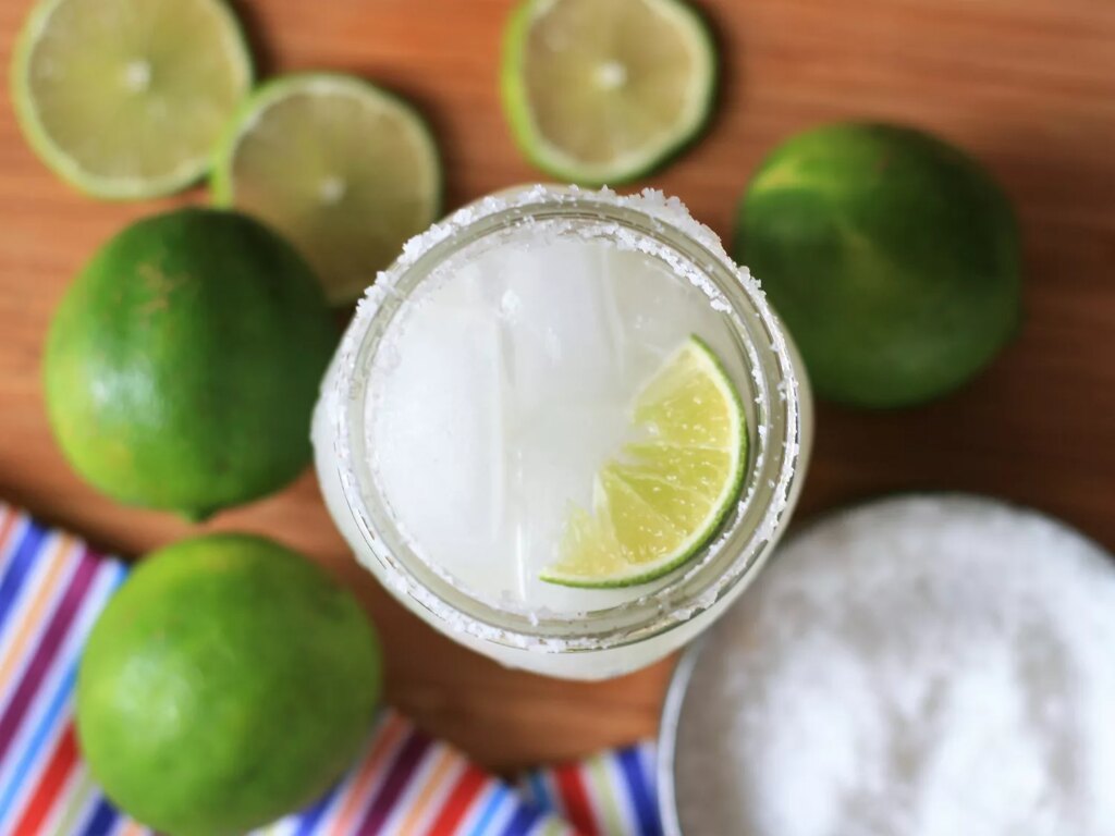 Overhead view of a keto margarita in a glass garnished with a lime wedge, with limes, lime slices and a bowl with salt in the background