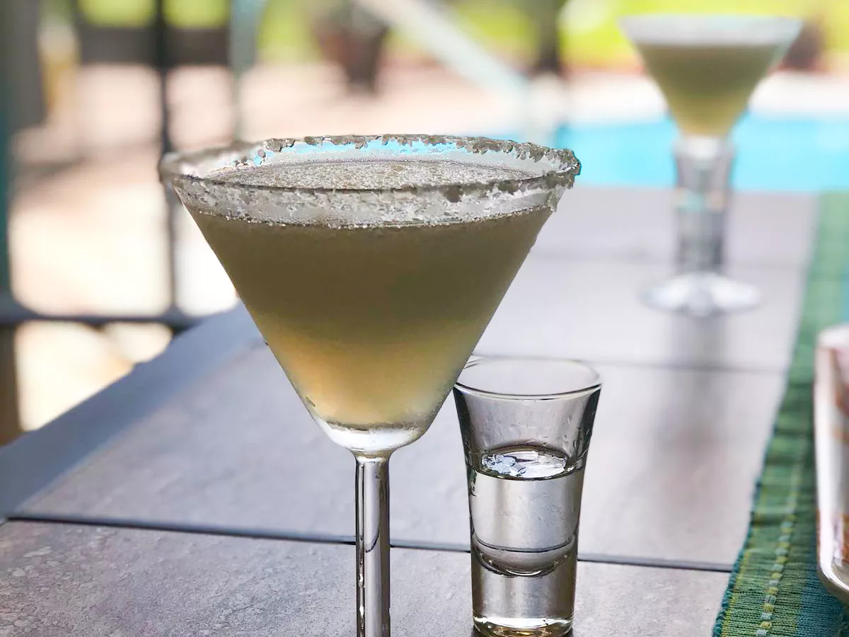 Close up view of a frozen margarita in a glass garnished with salt, next to a shot glass with clear liquid