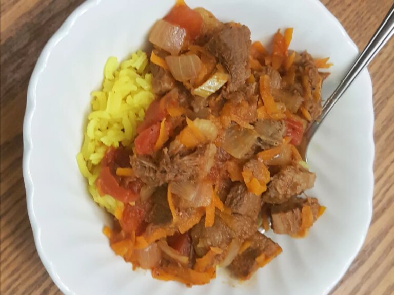Close up view of lengua (beef tongue) stew with carrots, tomatoes and onions, served over rice, on a white plate with a fork