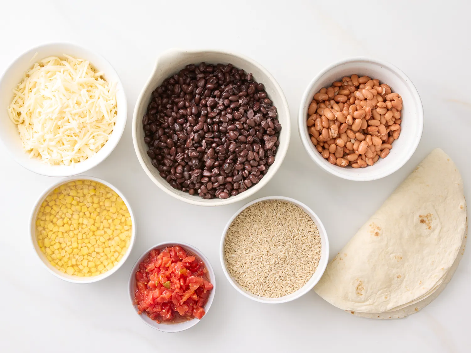 Bowls of shredded cheese black beans pinto beans corn kernels diced tomatoes quinoa and tortillas laid out