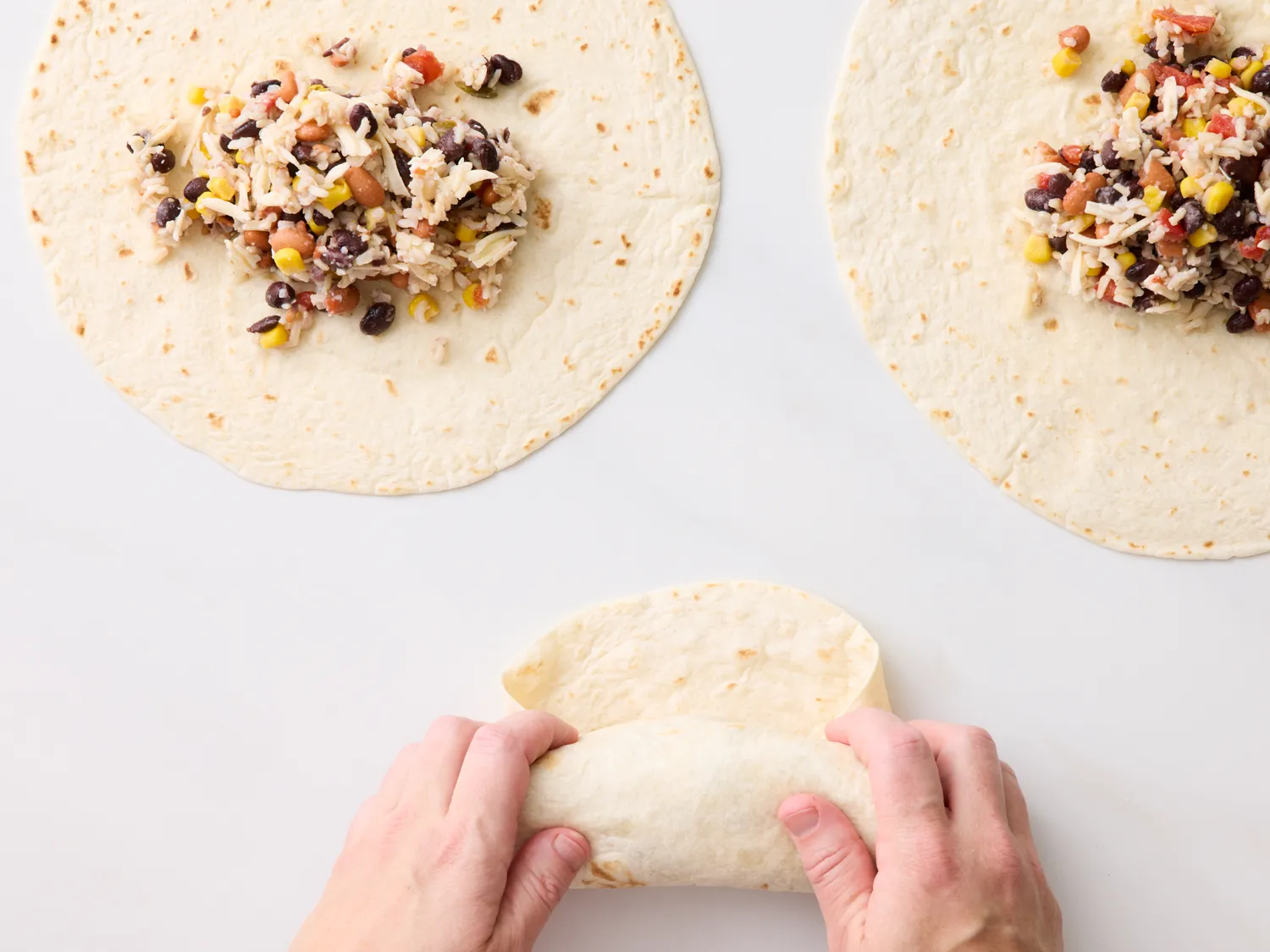 Hands rolling a tortilla with a filling of beans rice and vegetables with two other tortillas prepared on a white background