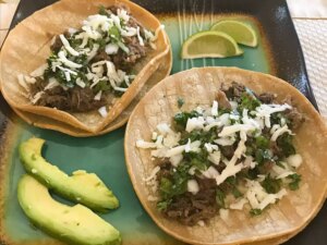Close up view of beef tongue tacos garnished with cheese and fresh herbs, served with lime wedges and avocado on a platter
