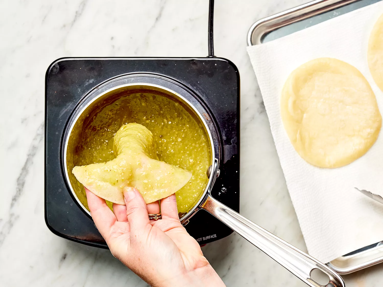 Hand dipping a corn tortilla into green salsa in a saucepan on a burner 