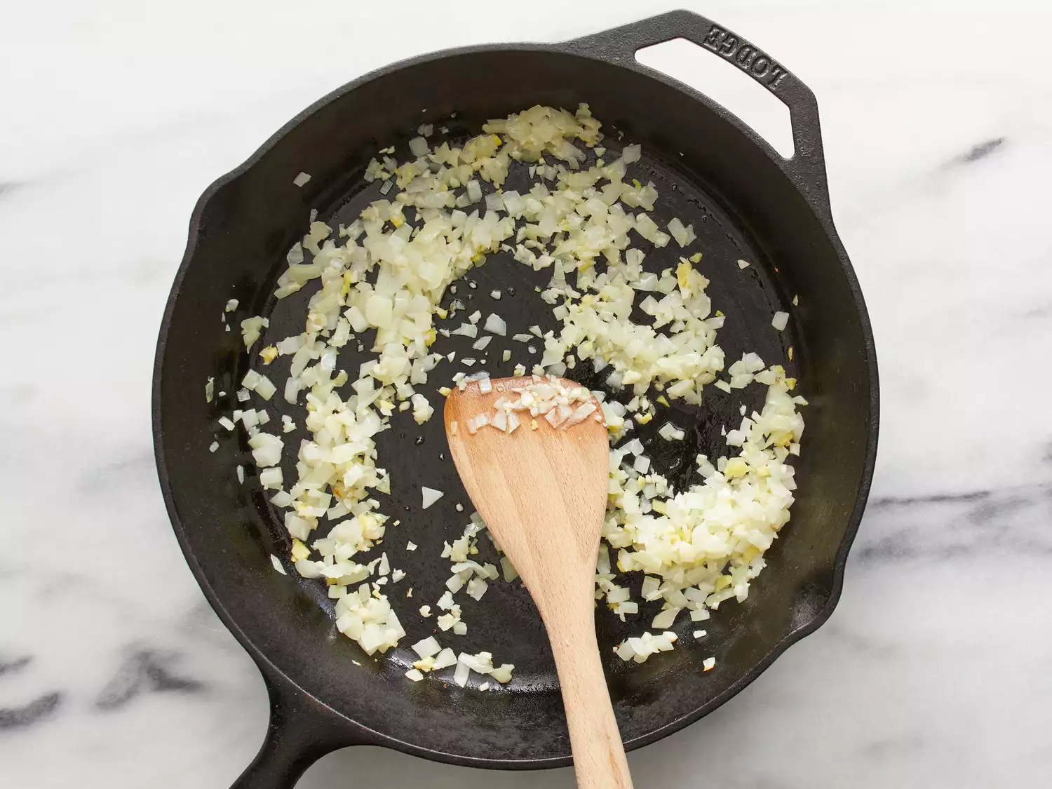 an overhead shot of onions, ginger, and garlic in a cast iron skillet with a wooden spoon