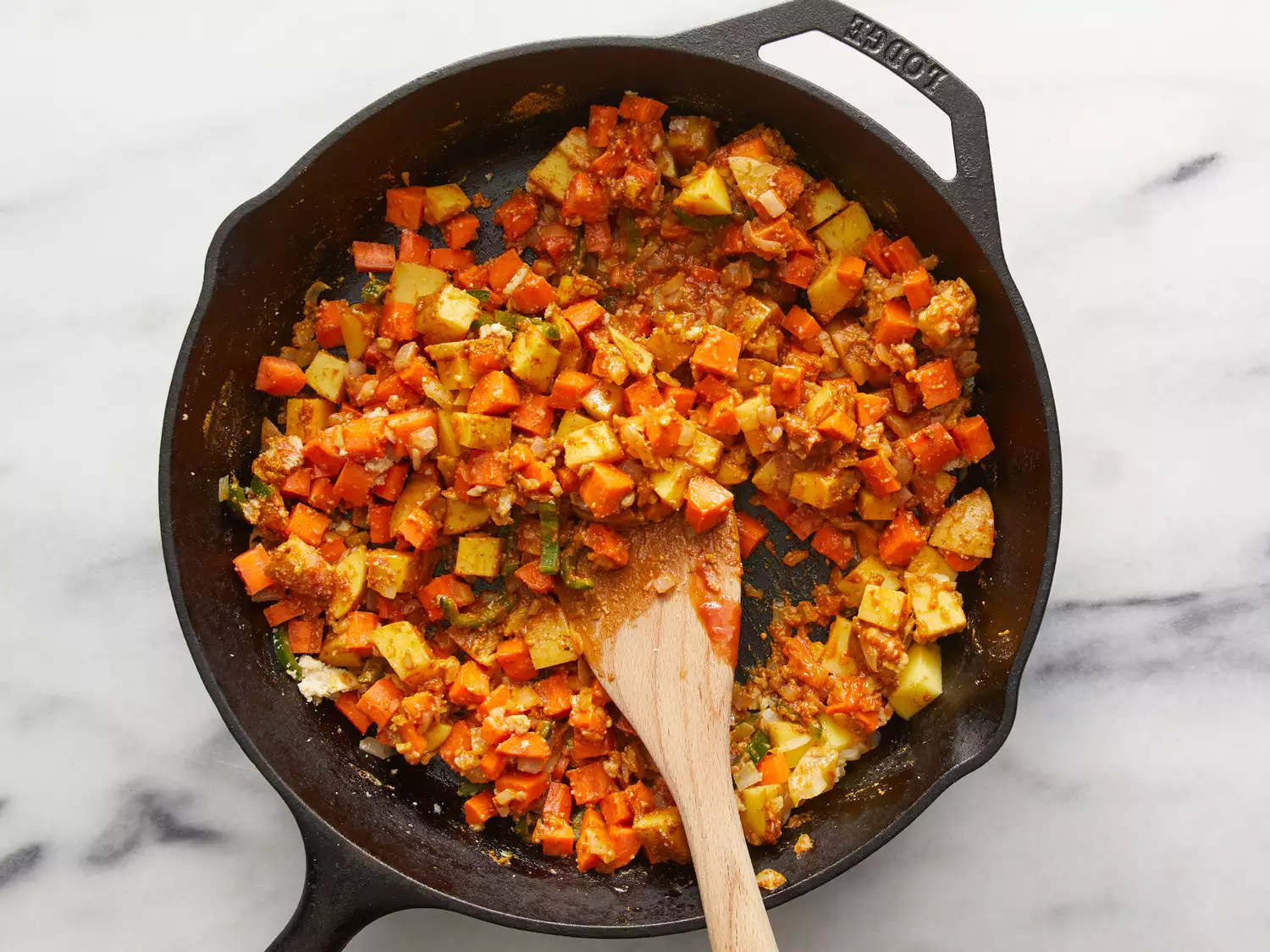 an overhead shot of carrots, potatoes, tomato sauce, cashews, and jalapeÃ±o added to a cast iron skillet with a wooden spoon