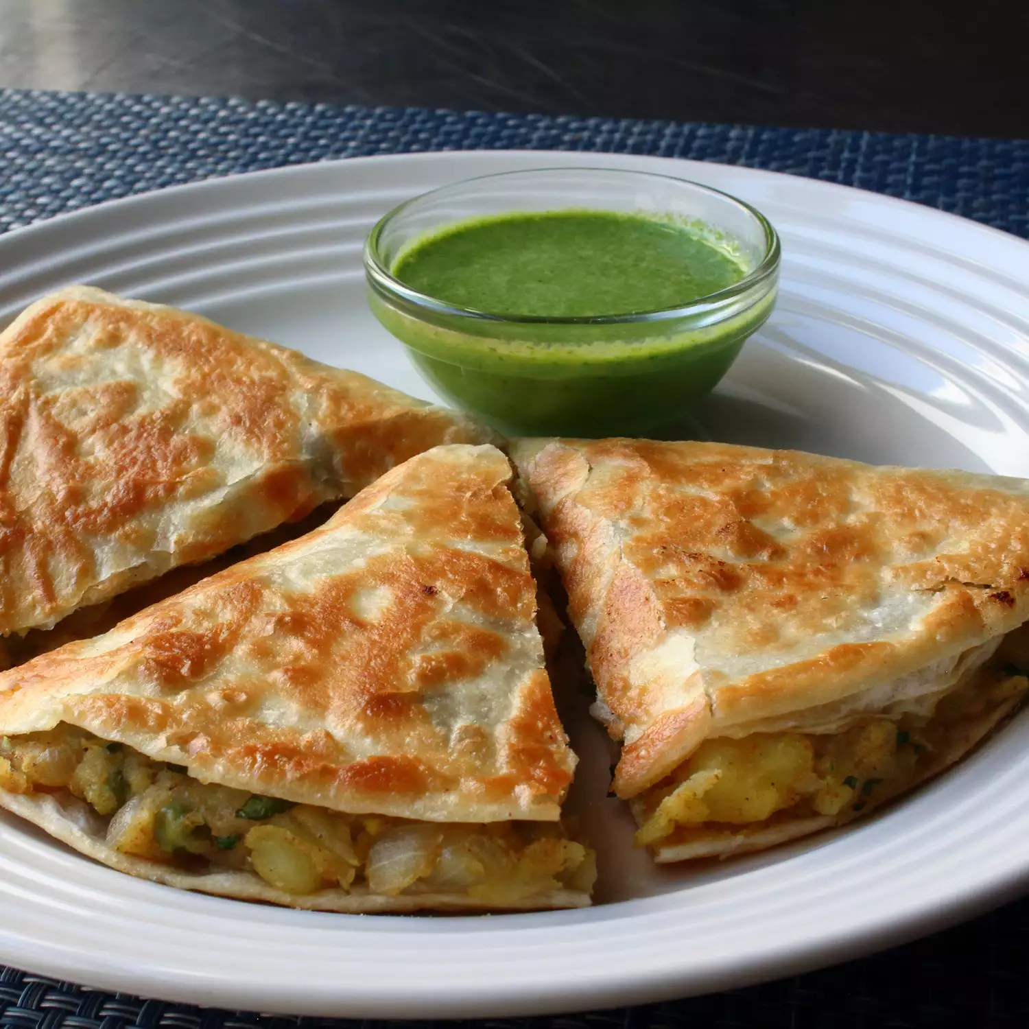 Close up view of a sliced samosadilla (samosa quesadilla) served with green sauce in a glass sauce cup on a white plate
