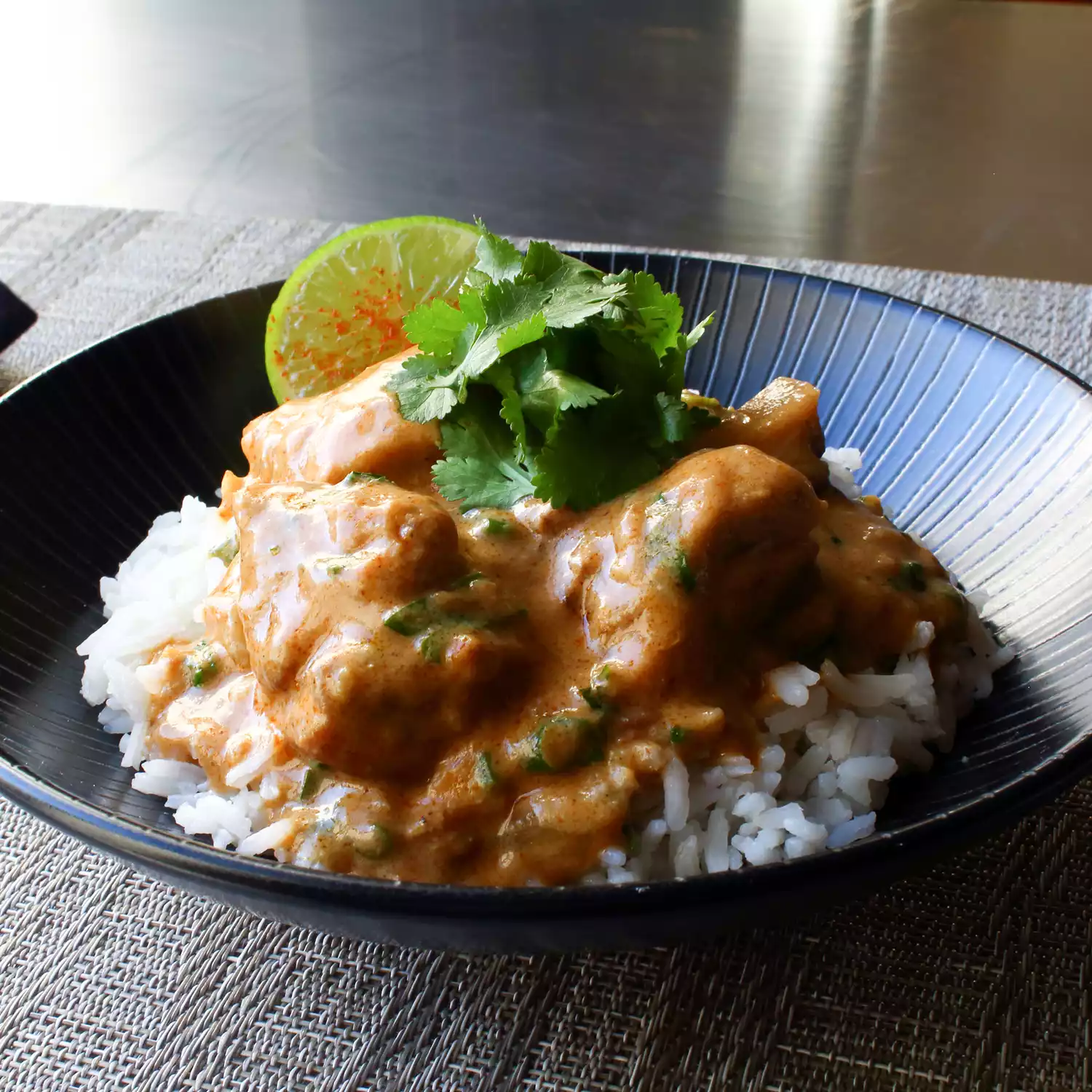 close up view of Creamy Cashew Chicken Curry over white rice, garnished with fresh herbs and a lime wedge in a black bowl